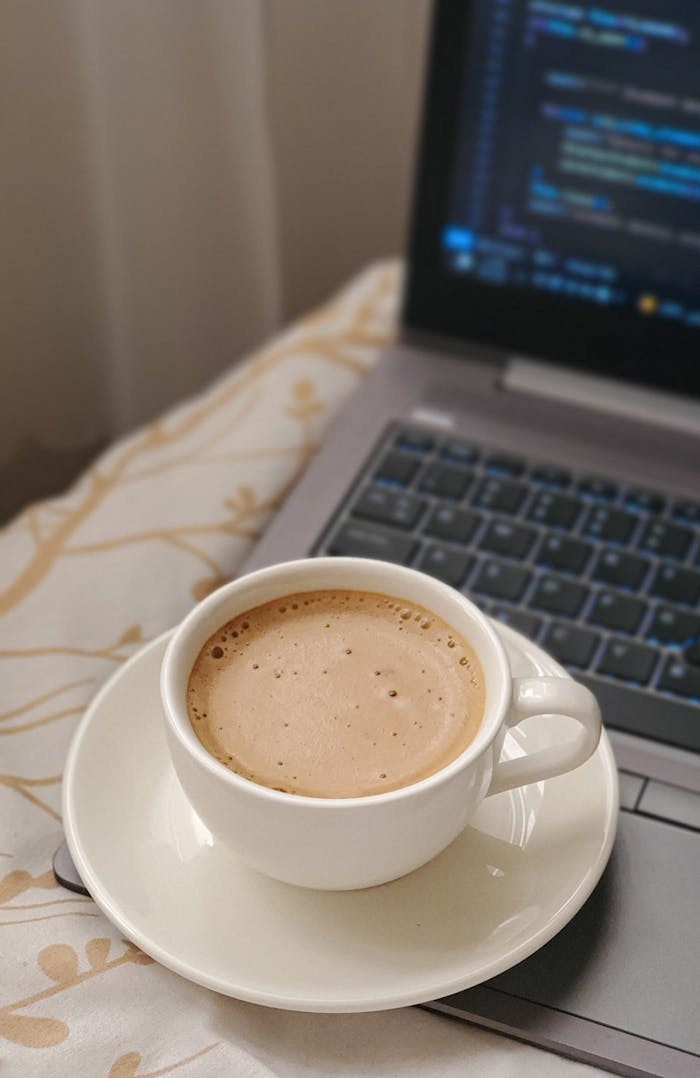 Home Cup of frothy coffee on a laptop keyboard with coding screen, cozy indoor setting.